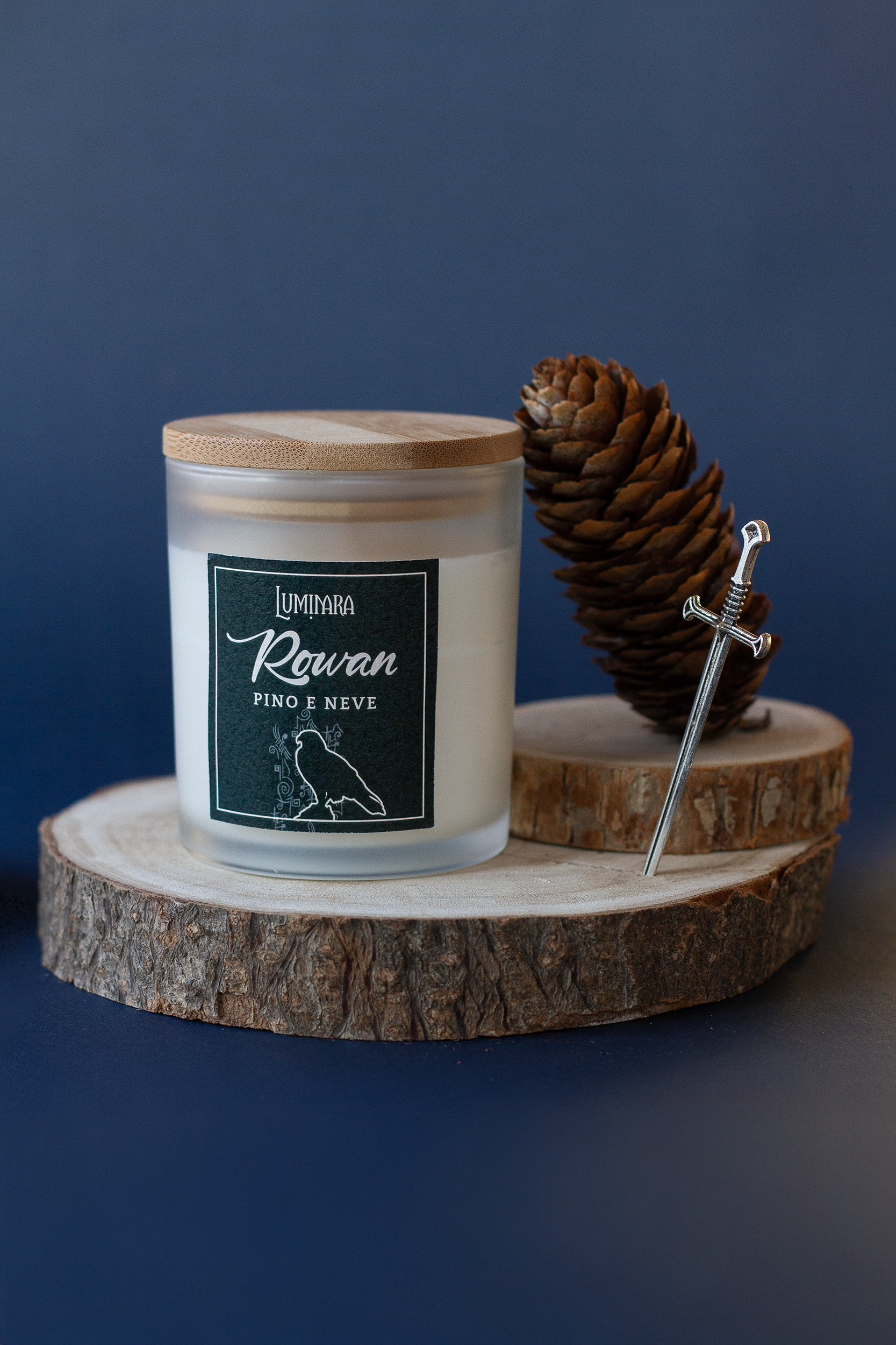 Candela elegante in vetro e tappo in bamboo ispirata a Rowan Whitethorn diLuminara Portali - etichetta vedre foresta effetto cuoio con scritto Rowan e  la profumazione pino e neve, ispirata a Terrasen e Trono di Vetro - Sarah J. Maas. Sullo sfondo una spada e una pigna