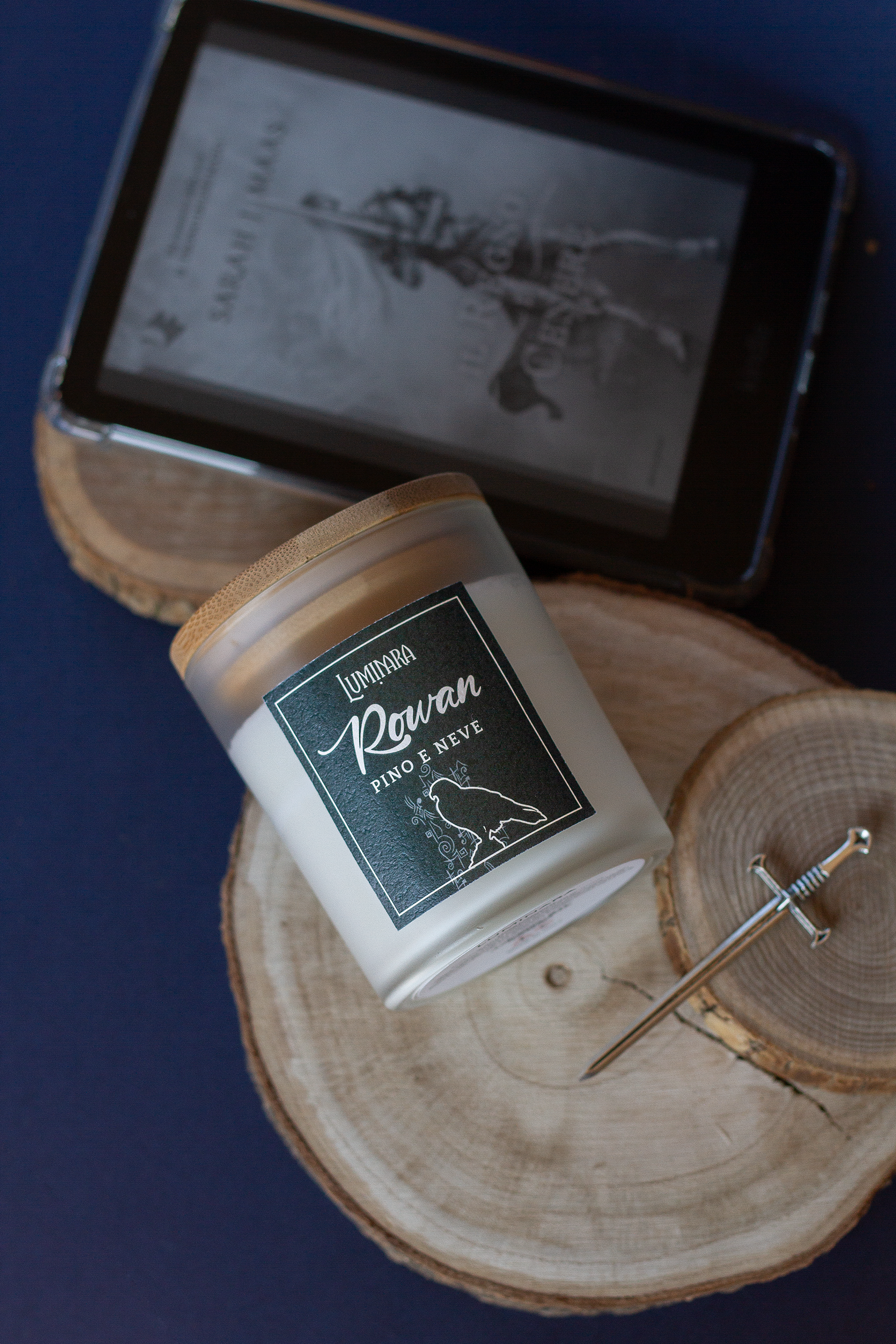 Candela elegante in vetro e tappo in bamboo ispirata a Rowan Whitethorn diLuminara Portali - etichetta vedre foresta effetto cuoio con scritto Rowan e  la profumazione pino e neve, ispirata a Terrasen e Trono di Vetro - Sarah J. Maas. Sullo sfondo una spada e un ereader con l'ebook Il regno di cenere