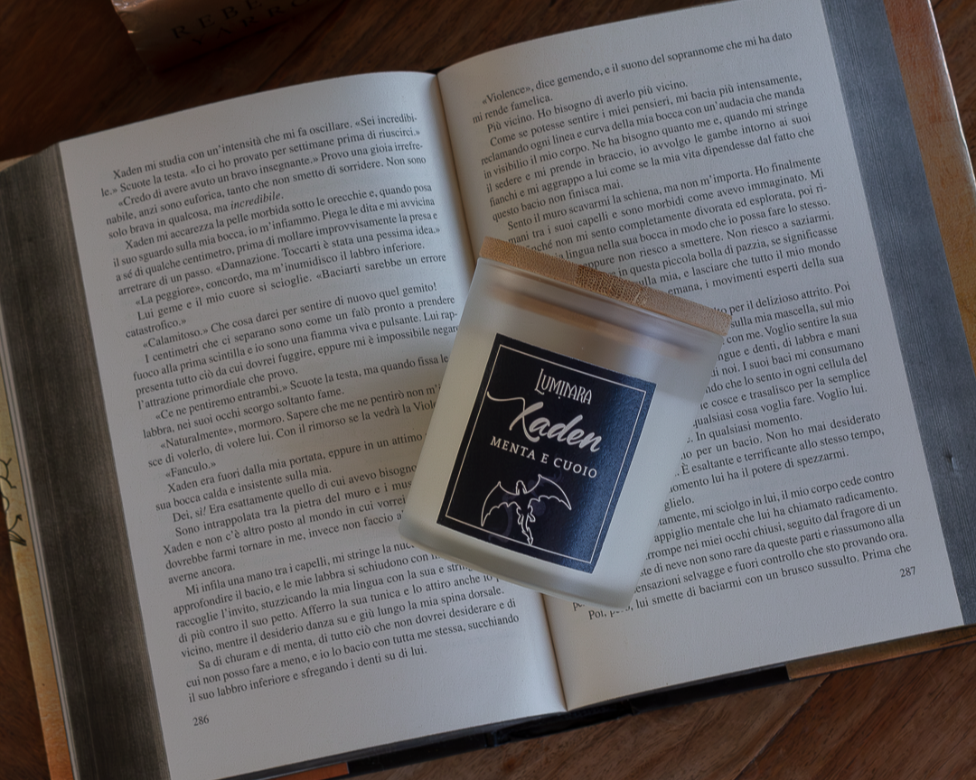 Candela elegante in vetro con tappo di bamboo, sull'etichetta è presente il nome Xaden e la profumazione menta e cuoio, ispirata a Fourth Wing e Iron Flame - Rebecca Yarros. Sullo sfondo il libro Fourth Wing aperto sulla scena del bacio tra Violet e Xaden