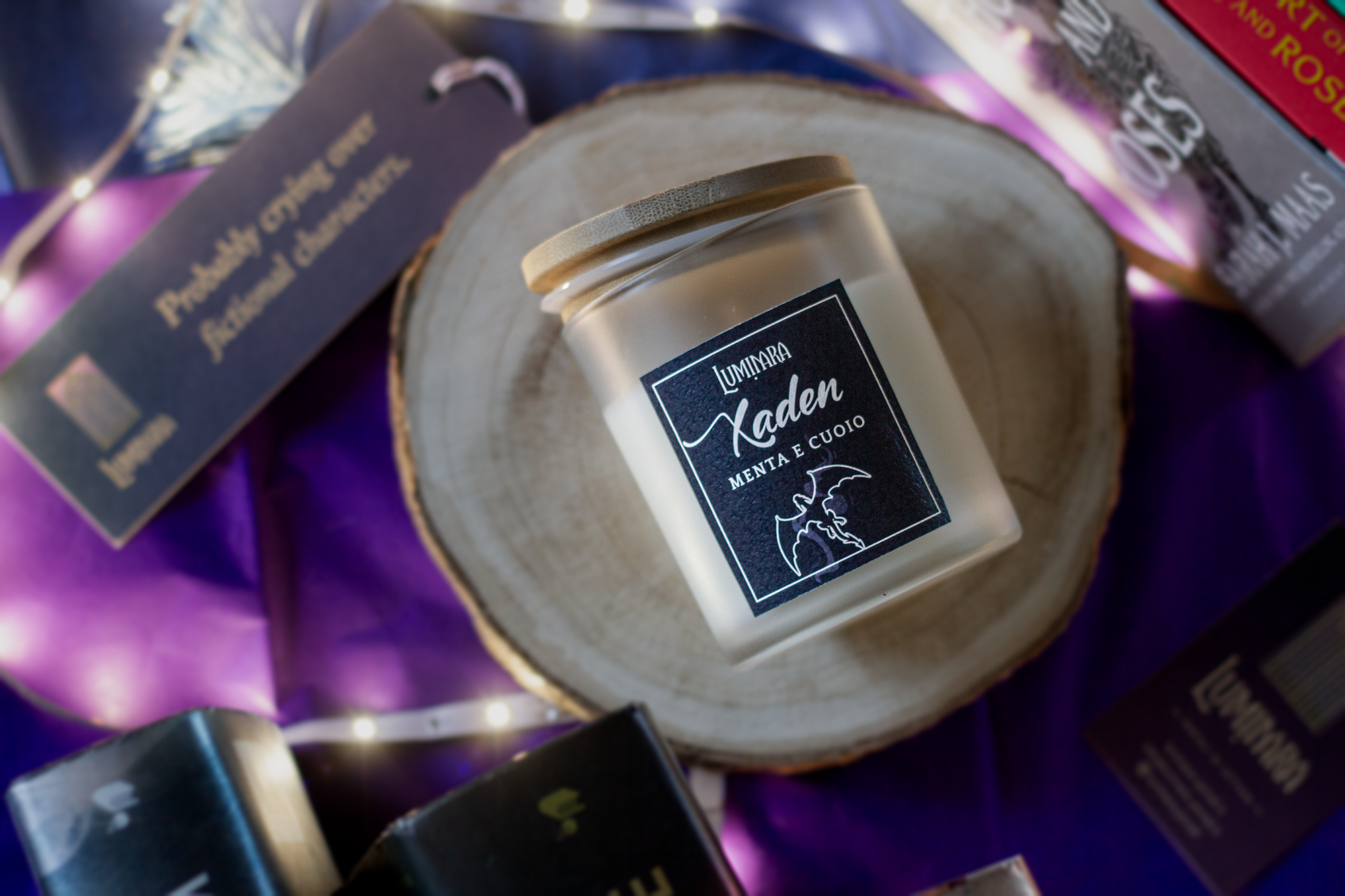 Candela elegante in vetro con tappo di bamboo, sull'etichetta è presente il nome Xaden e la profumazione menta e cuoio, ispirata a Fourth Wing e Iron Flame - Rebecca Yarros. Sullo sfondo libri ACOTAR e la serie Fourth Wing. Toni viola e luci. testo "probably crying over fictional characters" su un segnalibro
