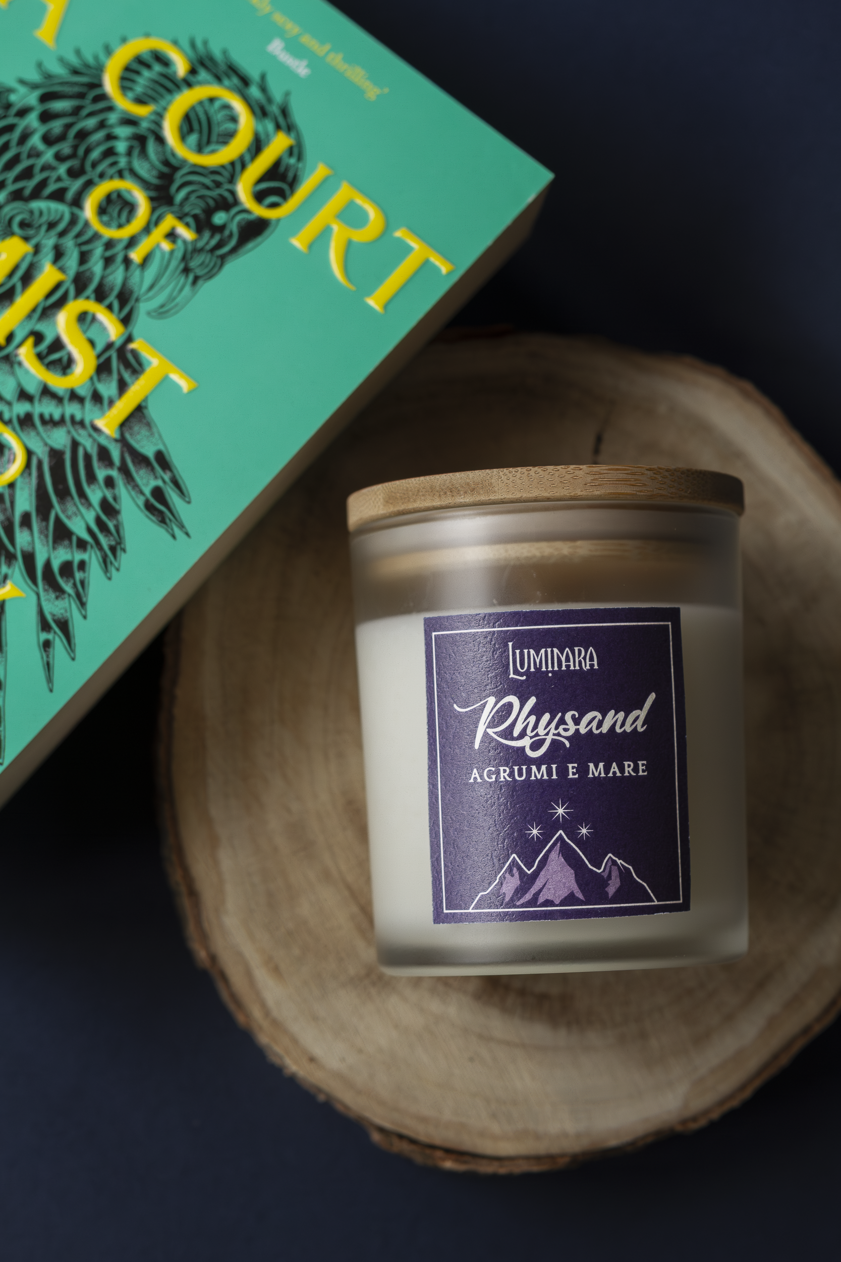Candela in vetro con tappo in bamboo, sull'etichetta il nome Rhysand di ACOTAR di Maas e la profumazione Agrumi e mare. Copertina del libro A court of mist and fury