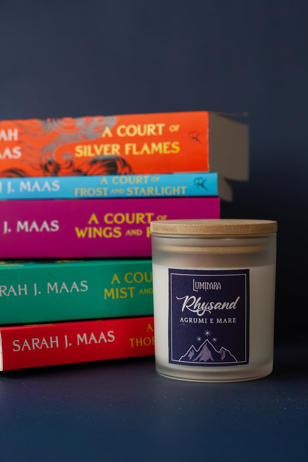 Candela in vetro con tappo in bamboo, sull'etichetta il nome Rhysand di ACOTAR di Maas e la profumazione Agrumi e mare. sullo sfondo la serie completa dei libri ACOTAR di Maas